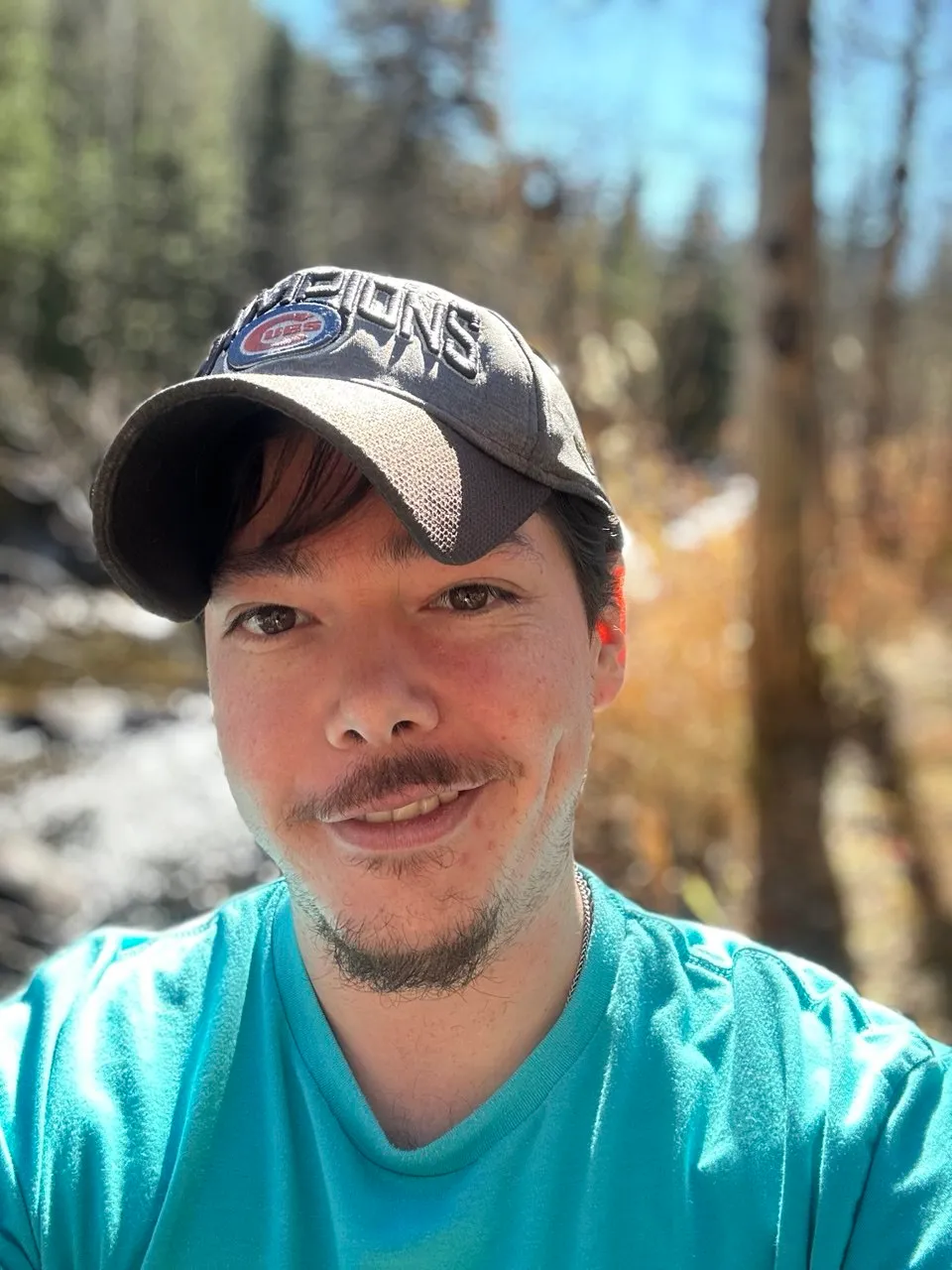 Sebastian in a cap, smiling, with a creek and trees behind him