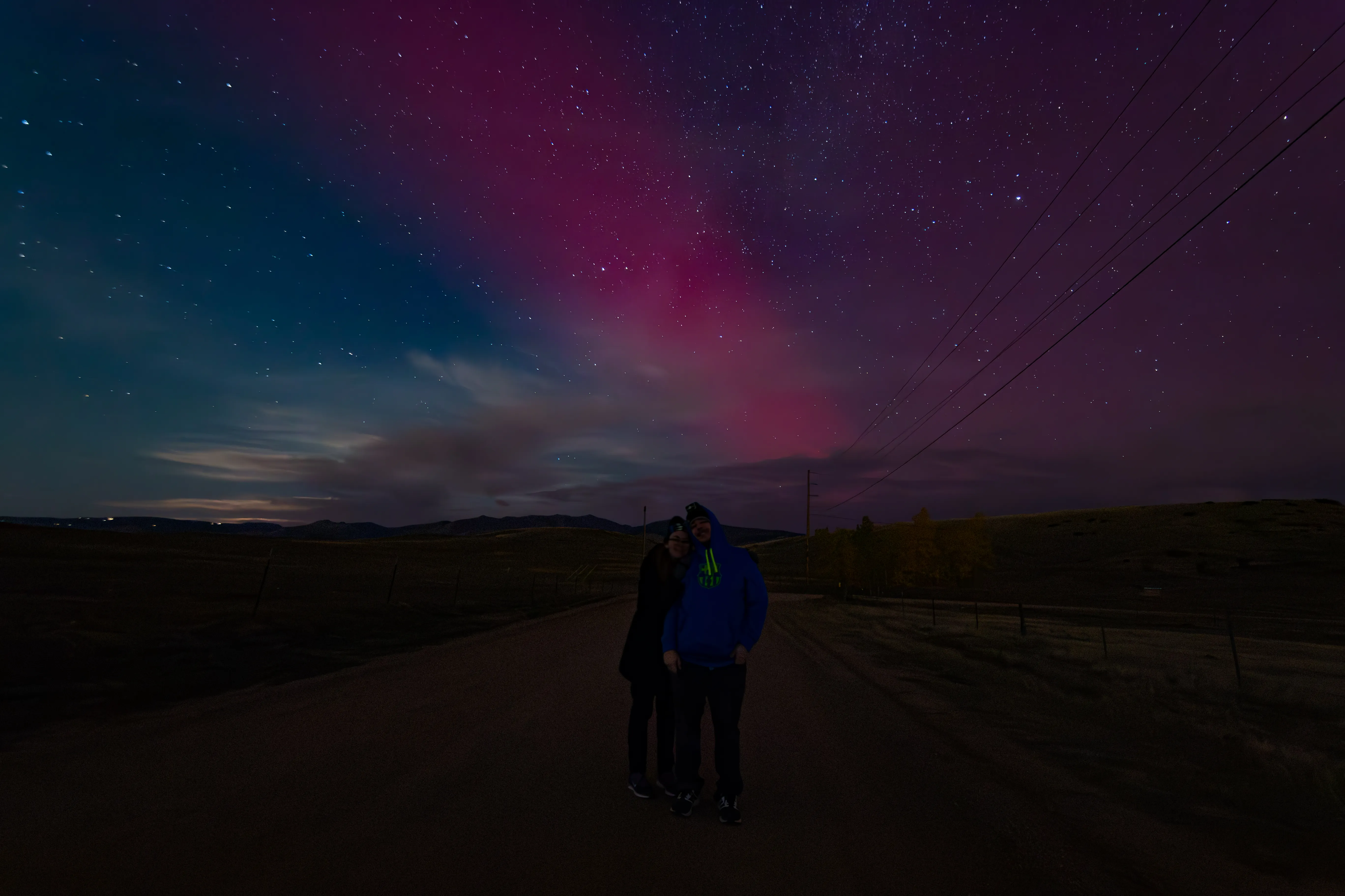 Pink and green aurora over a dark road at night, two people standing on the shoulder