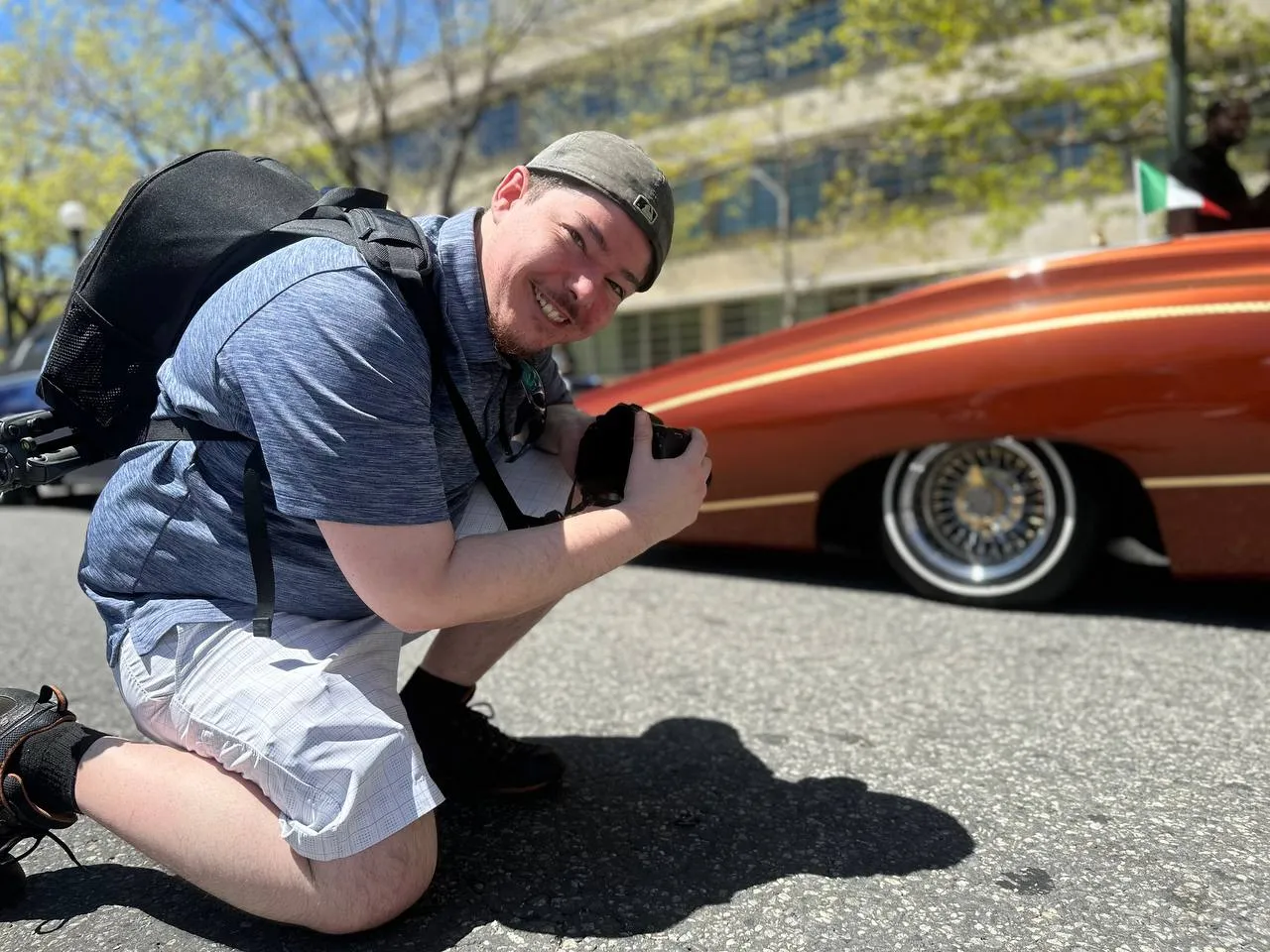 Sebastian kneeling with a camera beside a classic red car at a car show